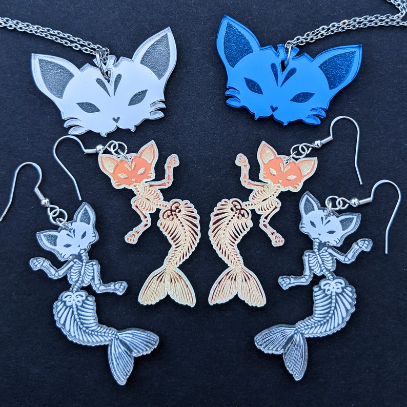 May include: A set of cat-themed jewelry featuring a white cat necklace, a blue cat necklace, and a pair of skeleton cat mermaid earrings. The earrings are available in two color variations: orange and silver.