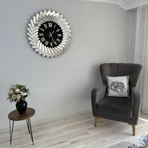 May include: A gray armchair with a white pillow featuring an elephant design sits in a room with a mirrored clock on the wall. The clock has a black face with Roman numerals and silver hands.