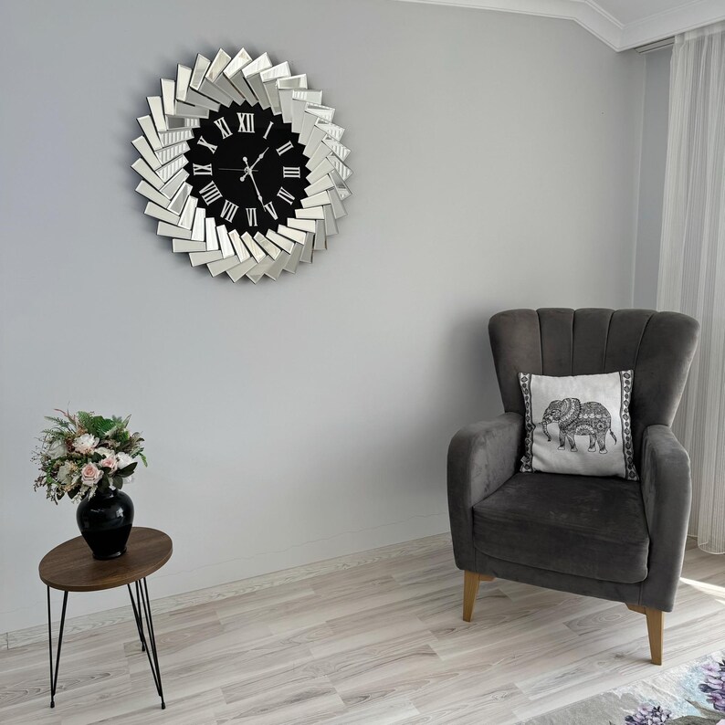 May include: A gray armchair with a white pillow featuring an elephant design sits in a room with a mirrored clock on the wall. The clock has a black face with Roman numerals and silver hands.