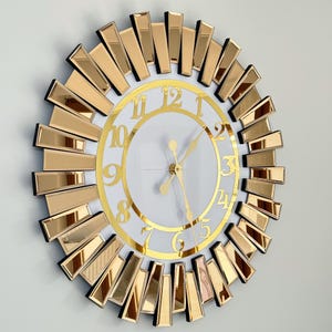 Peut inclure: Une horloge dorée et blanche avec un design en forme de soleil miroir. Le cadran de l'horloge comporte des chiffres romains et des aiguilles dorées.