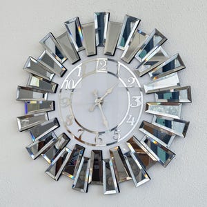 Può includere: Un orologio da parete decorativo con un design a raggiera a specchio. L'orologio ha un quadrante bianco con numeri e lancette argentate. Pezzi di specchio rettangolari circondano il quadrante, creando un effetto radioso. L'orologio è montato su una parete bianca.