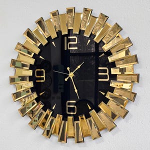 May include: A round, black wall clock with gold-colored rectangular accents radiating from the center. The clock face features gold hands and large gold numerals at the 12, 3, 6, and 9 o'clock positions. The clock has a modern, decorative design.