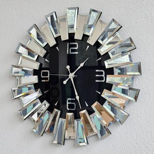 May include: A decorative wall clock with a black face and silver hands. The clock features large, mirrored rectangular accents around the perimeter, creating a sunburst effect. The numerals 3, 6, 9, and 12 are visible.