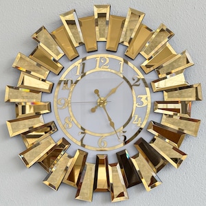 May include: A gold and white wall clock with a sunburst design. The clock features a white face with gold numerals and hands. The outer edge is adorned with rectangular gold mirrored accents, creating a decorative and elegant timepiece.
