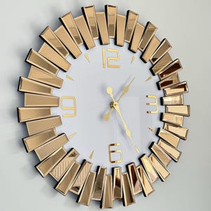 May include: A large, round, gold-toned wall clock with a white face and black Roman numeral hour markers. The clock is surrounded by a ring of mirrored, gold-toned, rectangular pieces.