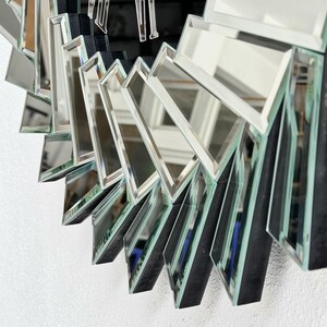 May include: Close-up of a mirrored wall decor with a sunburst design. The mirror is made up of many small, angled pieces of glass, each with a black border. The mirror has a modern and geometric design.
