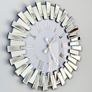 Może przedstawiać: Duży, okrągły, lustrzany zegar ścienny z rzymskimi cyframi godzinowymi i srebrnymi wskazówkami. Tarcza zegara jest biała, a płytki lustrzane są ułożone w kształcie słońca.