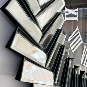 May include: A black and silver mirrored clock with Roman numeral markings. The clock face is made up of multiple mirrored pieces arranged in a sunburst pattern.