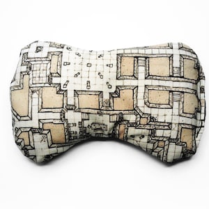 May include: A beige and brown pillow with a black and white dungeon map design. The pillow is shaped like a bow tie.