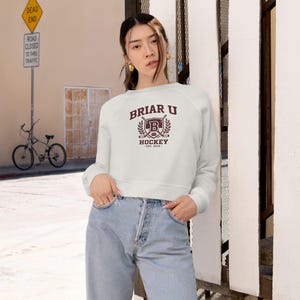 Puede incluir: Una sudadera corta blanca con el texto "Briar U Hockey" en granate. El diseño incluye un escudo con una "B" y palos de hockey cruzados. La sudadera se combina con vaqueros azul claro.