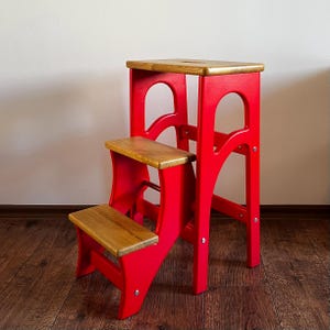May include: A two-step wooden step stool. The frame is painted red, and the steps are natural wood. The top step has a rectangular cutout. This step stool is designed for reaching higher places and is suitable for various household tasks.