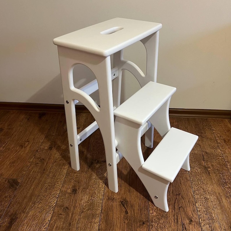 May include: A white wooden step stool with two steps and a top platform. The stool has a rectangular top with a handle cutout and curved supports. The steps are also rectangular, and the stool is painted white.