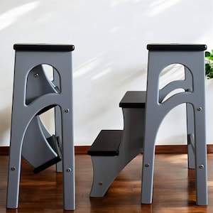 May include: Two-step wooden step stool in grey with black steps and top. The step stool has a unique arched design on the sides and is suitable for reaching higher shelves or cabinets. The step stool is made of wood and has a classic design.