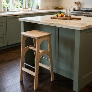 May include: A light-colored wooden kitchen stool with a square seat and arched side supports. It features a footrest and is set against a kitchen island with a marble countertop and green cabinets. The stool's design is simple and functional.