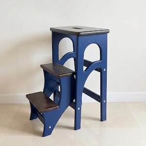 May include: A wooden step stool featuring a dark blue frame and three dark brown horizontal steps. The top step includes a rectangular cutout handle. The side panels have arched cutouts, and the structure is held together by visible metal screws.