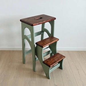 Puede incluir: Un taburete de madera con estructura de color verde salvia y tres peldaños de madera teñida en marrón oscuro. El peldaño superior incluye un asa recortada para transportarlo. La estructura tiene soportes laterales arqueados y una base firme.