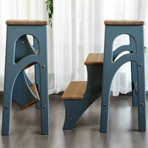 May include: Two blue step stools with wooden steps and tops. The stools have a unique arched design on the sides. The steps and tops are made of light-colored wood, contrasting with the blue frames.