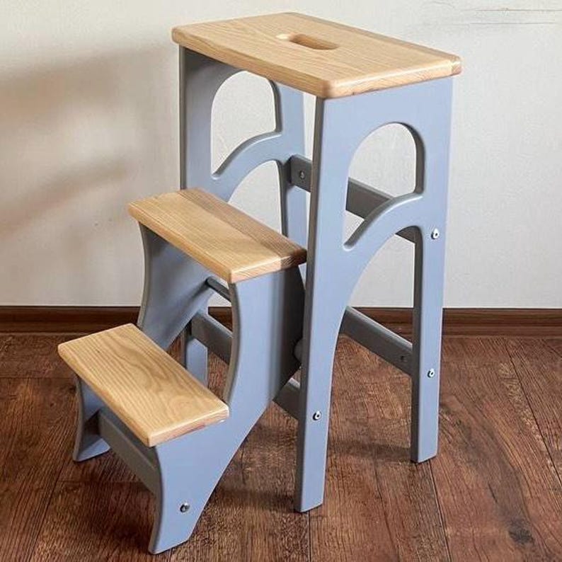 May include: A two-step wooden step stool with a light gray frame and natural wood steps. The top step has a handhold. The stool is designed for reaching higher places and is suitable for various household tasks.