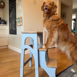 May include: A light grey wooden two-step stool featuring natural wood grain treads on the top and middle steps. The stool has a sturdy frame with arched side cutouts and visible metal hardware connecting the light grey painted side panels.