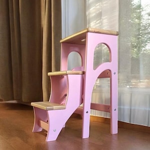 Op de afbeelding: Een roze houten opstapje met twee treden en een bovenste platform. De treden en het platform hebben een natuurlijke houten afwerking. De kruk heeft een boogvormig ontwerp aan de zijkanten en is ontworpen voor kinderen. De kruk is ongeveer 76 cm hoog.