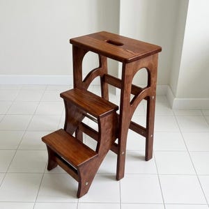 May include: A two-step wooden step stool with a rich brown finish. The stool has a rectangular top with a handle cutout and arched side supports. The lower step is also rectangular, and the design is simple and functional.