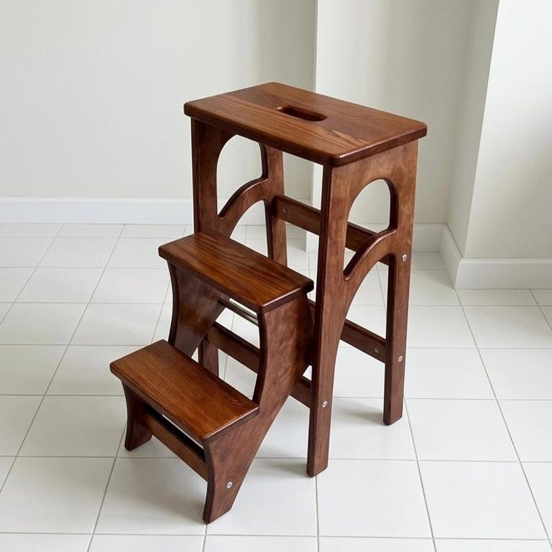 May include: A two-step wooden step stool with a rich brown finish. The stool has a rectangular top with a handle cutout and arched side supports. The lower step is also rectangular, and the design is simple and functional.