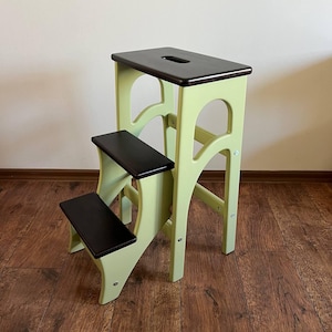 May include: A two-step wooden step stool. The frame is painted light green, and the steps are black. The top step features a handle. This step stool is designed to help reach higher areas and is suitable for use around the home.