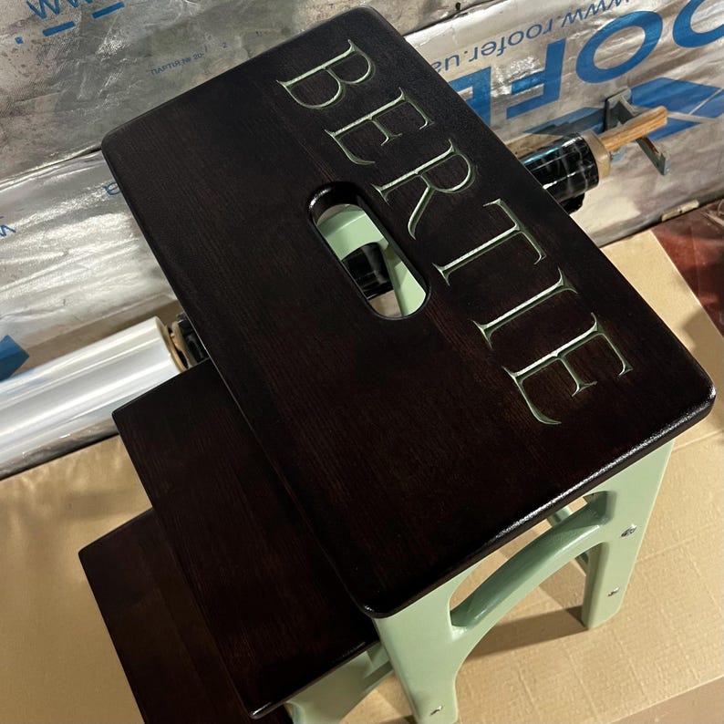May include: A dark brown wooden step stool with the name "BERTIE" carved into the top step. The legs and supports are a light green color. The stool has a handle cut out of the top step.
