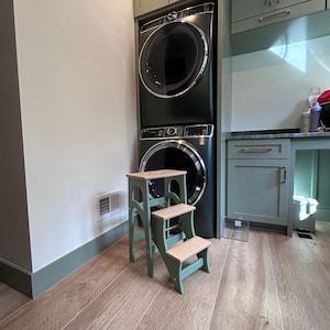 May include: A small, sage green wooden step stool with three steps and natural wood-colored treads. The stool is positioned in front of a dark green stacked washer and dryer unit, with a light-colored wall and wood flooring.