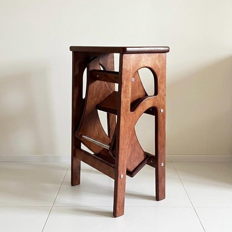 May include: A wooden step stool with a rich brown finish. The stool features a folding design with two steps and a safety bar. The frame has cut-out details and is supported by four legs. The stool is designed for use in the home.