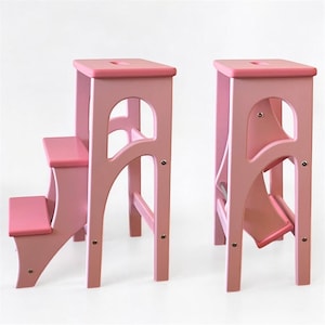 Puede incluir: Dos taburetes de madera rosa. Uno tiene dos escalones, el otro un solo escalón plegable. Ambos tienen tapas cuadradas con un pequeño recorte y soportes laterales arqueados. Los taburetes están pintados de color rosa claro. Altura aproximada 60 cm.