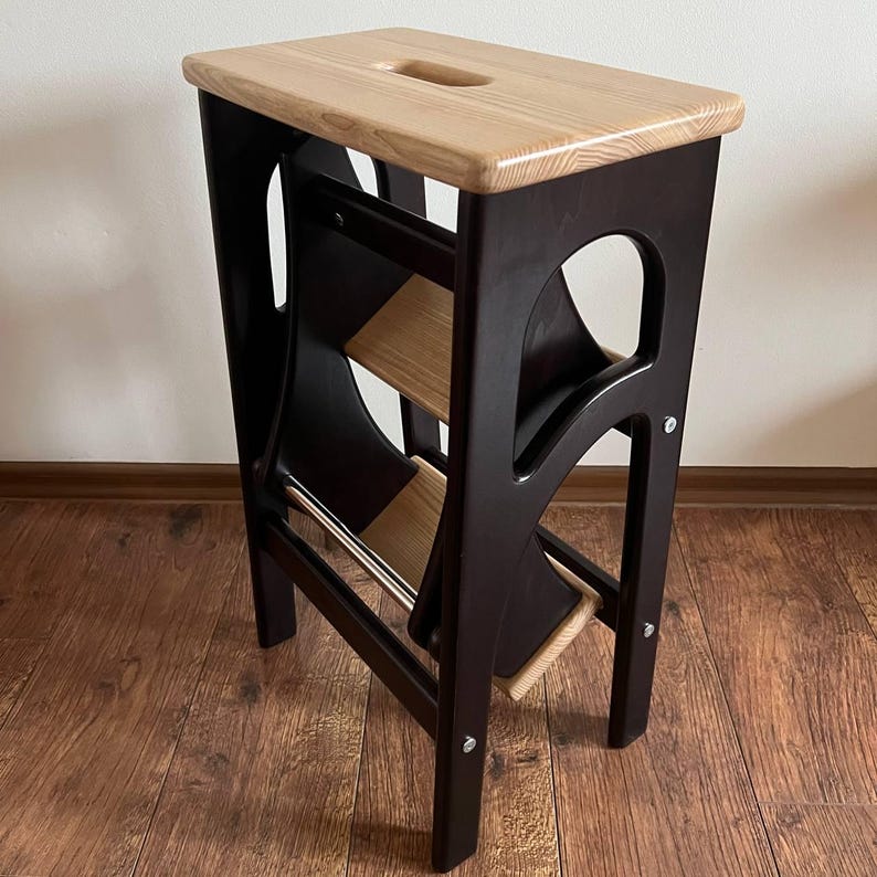 May include: A wooden step stool with a dark brown frame and natural wood steps and top. The top has a handle cutout. The stool is designed to fold for storage. The steps are made of light-colored wood, contrasting with the darker frame.