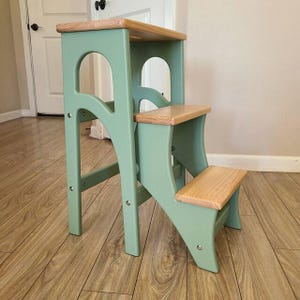Puede incluir: Taburete de dos escalones de madera, con estructura pintada de verde claro y escalones y parte superior de madera natural. El taburete tiene un diseño arqueado en los laterales, adecuado para alcanzar lugares altos.