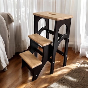 Puede incluir: Un taburete de madera con dos escalones y una plataforma superior. Los escalones y la parte superior son de madera de color claro, mientras que el marco es de color marrón oscuro. La plataforma superior tiene un recorte de asa. El taburete está diseñado para alcanzar lugares más altos.