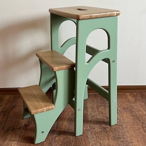 May include: A two-step wooden step stool with a light green frame and natural wood steps and top. The top step has a small, rectangular cutout. The stool is designed with arched supports and visible screw details, suitable for home use.
