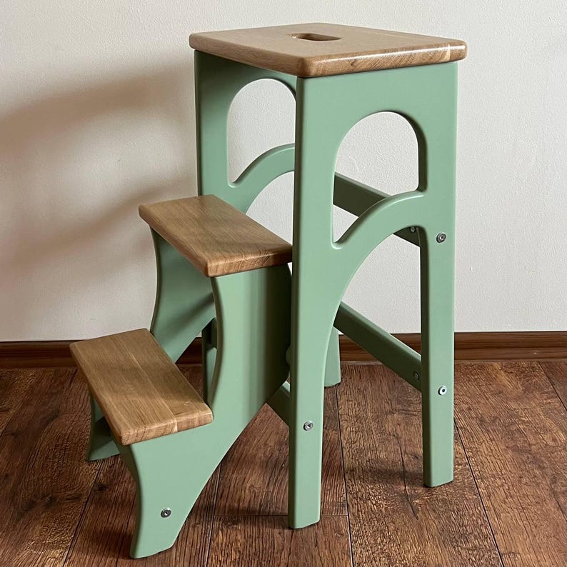 May include: A two-step wooden step stool with a light green frame and natural wood steps and top. The top step has a small, rectangular cutout. The stool is designed with arched supports and visible screw details, suitable for home use.