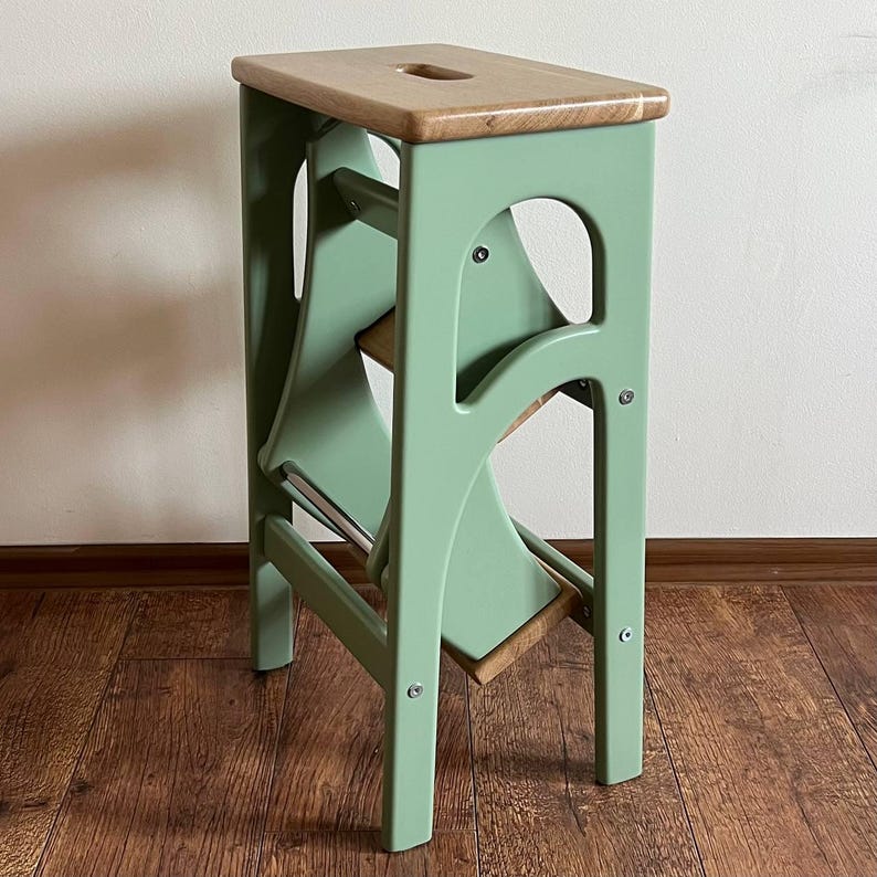 May include: A folding step stool with a light green frame and a natural wood top. The stool has a cut-out handle and two folding steps. The frame is painted a soft green, and the steps and top are made of natural wood.