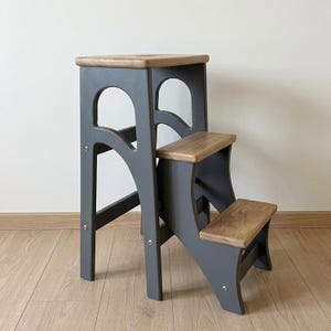 May include: A three-step wooden step stool featuring a dark grey painted frame with natural wood grain treads and a top platform. The structure includes arched side cutouts and is assembled with visible silver metal hardware screws.