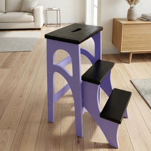 Puede incluir: Una escalera de mano de madera de tres peldaños con un marco de color violeta claro y peldaños horizontales de color marrón oscuro, casi negro. El peldaño superior incluye un asa rectangular recortada y la estructura se apoya en paneles laterales arqueados.