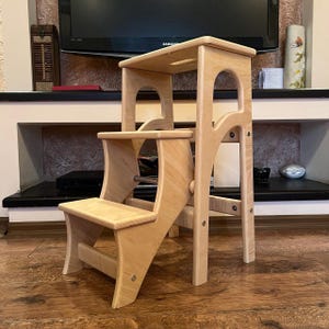 May include: A light-colored wooden step stool with three steps. The stool has a rectangular top step with a handle cutout, a middle step, and a bottom step. The frame features arched cutouts and is constructed with visible screws.
