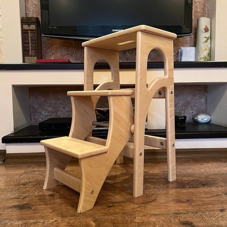 May include: A light-colored wooden step stool with three steps. The stool has a rectangular top step with a handle cutout, a middle step, and a bottom step. The frame features arched cutouts and is constructed with visible screws.