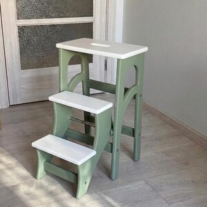 Op de afbeelding: Een houten opstapje met drie treden, voorzien van een saliegroen frame en witte treden. De bovenste trede heeft een rechthoekige uitsparing als handvat en de constructie wordt ondersteund door gebogen zijpanelen met een metalen stang.