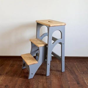 Könnte beinhalten: Ein dreistufiger Trittschemel aus Holz mit einer hellen Naturholzoberfläche auf den Stufen und der oberen Plattform, kontrastiert durch einen massiven, grau lackierten Rahmen. Die obere Plattform verfügt über einen Griffausschnitt.