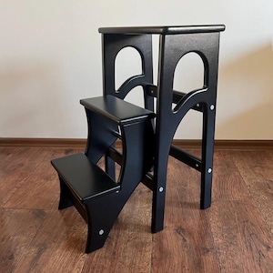 Puede incluir: Un taburete de madera negro con dos escalones y una plataforma superior. El taburete tiene un diseño clásico con soportes curvos y un acabado liso y brillante. Diseñado para alcanzar estantes o superficies más altas.