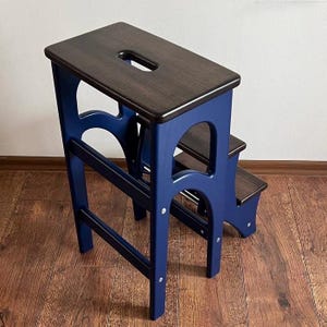 Op de afbeelding: Een houten opstapje met twee treden. Het heeft een donkerbruine bovenste trede en treden, en een marineblauw frame. De bovenste trede heeft een handgreepuitsparing. De kruk is ontworpen om hogere plaatsen te bereiken.