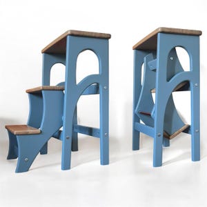 May include: Two wooden step stools featuring a blue frame and natural wood-toned steps. One stool is shown in an open, functional position with three tiers, while the other is folded into a compact, upright configuration with visible metal hardware.
