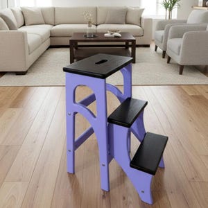 May include: A three-step wooden step stool. The frame is lavender, and the steps are black. The top step has a handle. The stool is designed for reaching higher shelves or cabinets. The step stool is made of wood.