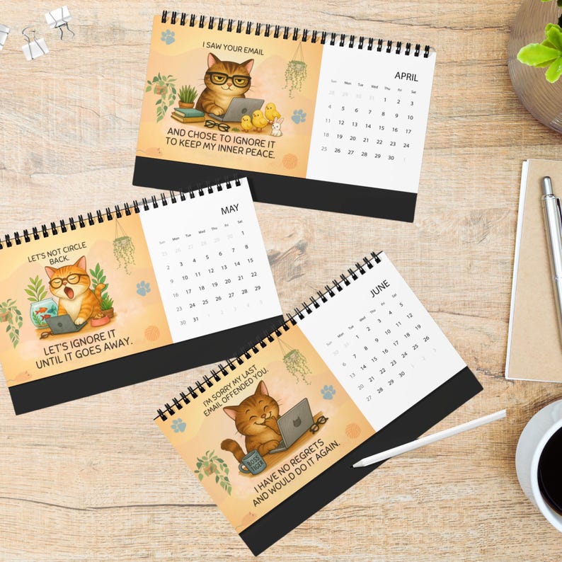 Op de afbeelding: Drie spiraalgebonden bureaukalenders met cartoon-katillustraties en humoristische kantoorteksten. Elke pagina toont een maand, waaronder april, mei en juni, met datums en afbeeldingen van katten met laptops, planten en koffiemokken.
