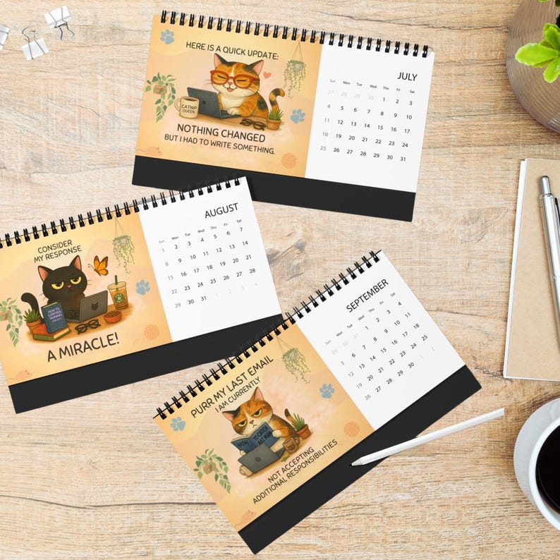 Op de afbeelding: Drie spiraalgebonden bureaukalenders met cartoon-katillustraties voor juli, augustus en september. Elke pagina toont een maandoverzicht, een kat die op een laptop werkt en grappige teksten zoals "Nothing changed but I had to write something."