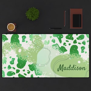 Pode incluir: Um tapete de mesa retangular com um design abstrato verde e branco. O tapete apresenta várias tonalidades de manchas verdes e o nome "Maddison" em uma fonte de script. Uma xícara de café, um caderno, uma caneta e uma planta estão em uma superfície preta.